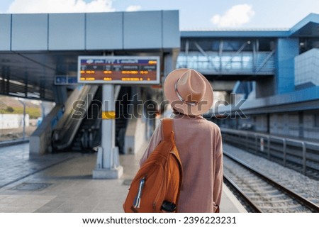 traveling by train. A young woman in a coat and hat stands at the station and looks at the train schedule. adventure and travel. Royalty-Free Stock Photo #2396223231