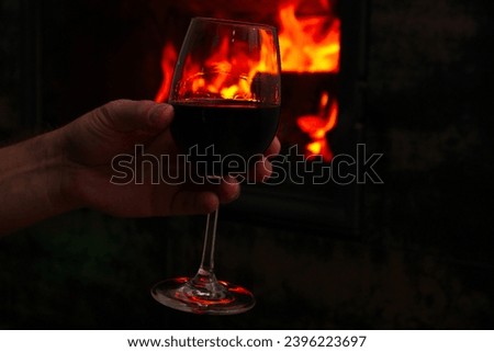 Mans hand holds the wine glass in front of the fireplace. Man with glass of wine near fireplace at home. Blur burning fireplace background.