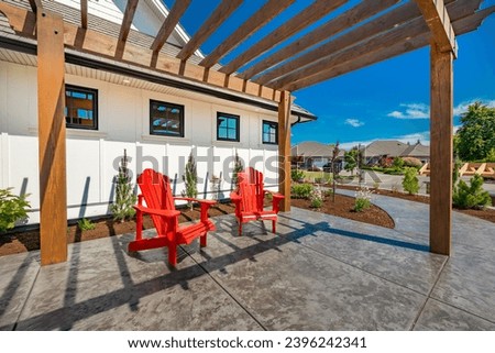 bright and sunny patio with pergola stamped concrete with two red Adirondack chairs  deep and beautiful blue sky bark mulch new landscaping a backdrop of a white home with windows reflecting the sky
