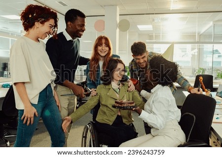 An interracial businesswoman is having a birthday surprise at the office with colleagues. A happy colleague with disability is about to hug her while a birthday girl is holding a cake. Bday at office.