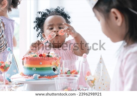 Selective focus on hand of African little child girl holding jelly, enjoy eating rainbow cake on birthday party with fork. Kids have fun celebrating party at home with friends