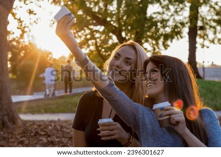 Two attractive young women taking selfie in the public park during sunset and refreshing with the coffee