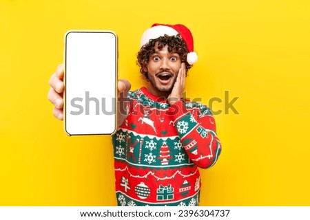 young shocked hindu man in new year clothes showing blank smartphone screen on yellow isolated background, surprised hindu guy in santa hat advertising mobile display online in amazement