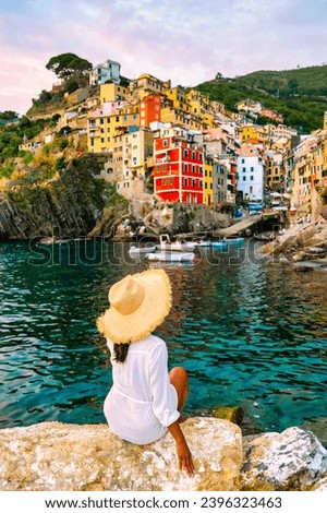 Riomaggiore Cinque Terre Italy, colorful village harbor front by the ocean, young Asian woman with a hat watching the sunset at waterfront looking out over Riomaggiore village Cinque Terre Italy Royalty-Free Stock Photo #2396323463
