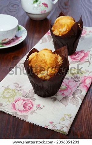 Muffins on a napkin on a wooden table. 2 plain cupcakes in butter paper on a floral napkin on a dark wooden surface with a tea set in the background, vertical image.