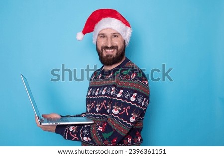 Merry young fun bearded IT man wear warm knitted sweater Santa hat posing hold use work on laptop pc computer on blue background. New Year 2024 holiday celebration concept. 
