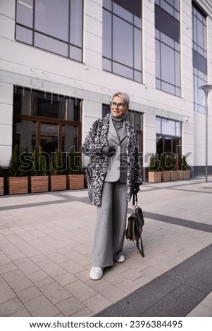 Stylish cool senior woman model in fashionable winter multilayer outfit, animal print jacket coat, grey wool suit with pants and turtleneck stands full length in city. Trending female in warm clothes.