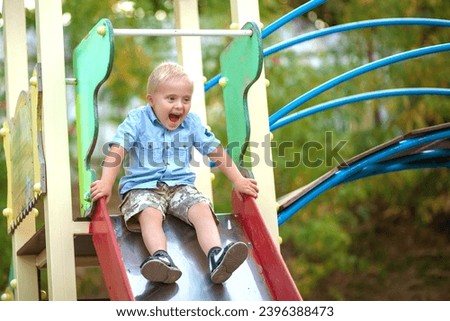 A little cute boy with Down syndrome rides on a slide on the playground. A child plays outside in a park in a play area.