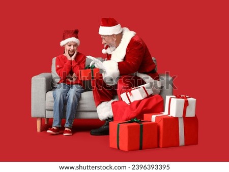 Santa Claus and cute little girl with gift boxes sitting on sofa against red background