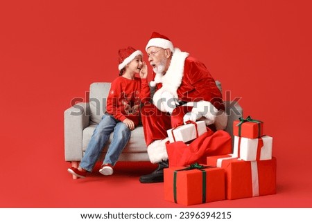 Shocked Santa Claus and cute little girl with gift boxes telling her wish on sofa against red background