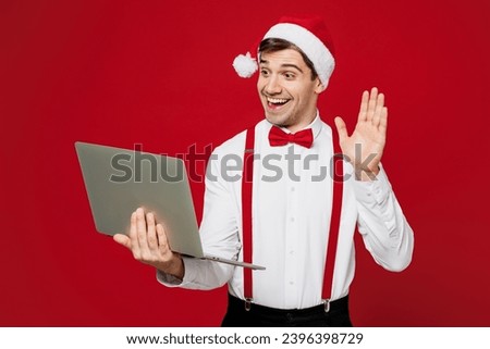 Merry young IT man wear white shirt Santa hat posing holding use work on laptop pc computer waving hand isolated on plain red background studio. Happy New Year Christmas celebration holiday concept