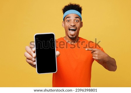 Young fitness trainer man sportsman wear orange t-shirt hold use point on blank screen area mobile cell phone spend time in home gym isolated on plain yellow background. Workout sport fit abs concept Royalty-Free Stock Photo #2396398801