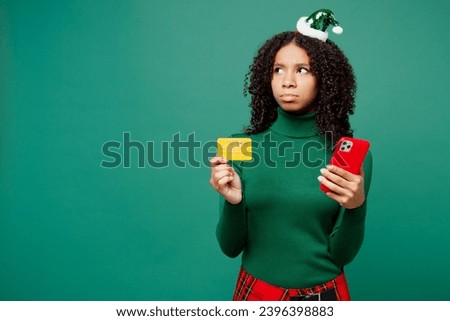 Merry little kid teen girl wears hat casual clothes posing using mobile cell phone credit bank card shopping online isolated on plain green background studio. Happy New Year Christmas holiday concept