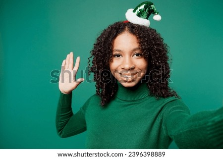 Merry little kid teen girl wear turtleneck hat casual clothes posing do selfie shot on mobile cell phone waving hand isolated on plain green background studio portrait. Happy New Year holiday concept