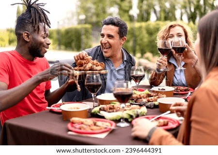 Friends of diverse ethnicities share a laugh over a sumptuous outdoor meal food, toasting with glasses of red wine. Royalty-Free Stock Photo #2396444391