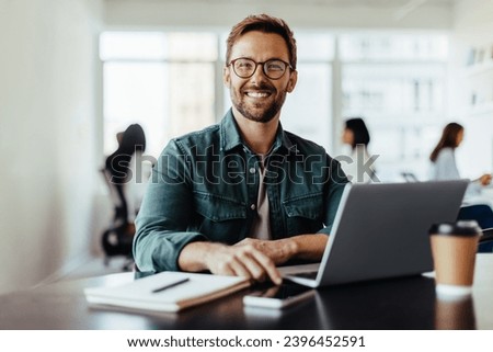 Portrait of a business man sitting in an office with his colleagues in the background. Happy business man working in a co-working office.