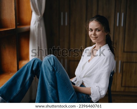 Woman sitting at home by a wooden window with a smile on a chair in homemade comfortable clothes and looking at the landscape, spring mood, women's day, rest on the weekend. Royalty-Free Stock Photo #2396460761
