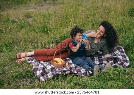 Cute little boy sitting on plaid with mom on picnic holds phone shows to mom pictures on screen. Family leisure, motherhood. Young woman enjoying vacation with son.