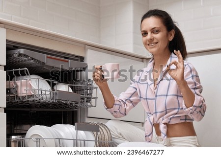 attractive woman unloading cup from open automatic built-in dishwasher machine and showing ok gesture in modern white kitchen.