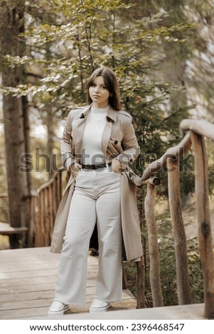 Vertical photo. Beautiful carefree young teenage girl in brown coat and white jeans and white sweater. Portrait of a beautiful girl in a brown raincoat against the background of the forest and nature.