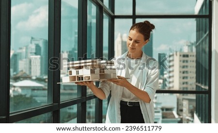 Young smart caucasian beautiful architect engineer raised house model while inspect carefully surrounded by city skyscraper view. Civil engineering, architectural studio. project planing. Tracery