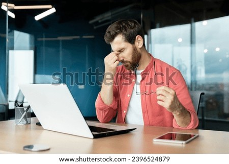 Sick man at workplace has eye pain and dizziness, businessman overworked and overtired working late with laptop sitting at desk inside office. Royalty-Free Stock Photo #2396524879