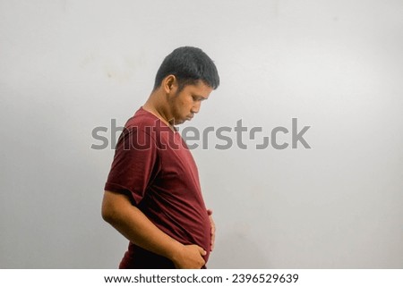 Portrait of handsome Asian man looking sideways in red t-shirt, looking at his bulging stomach on an isolated white background.