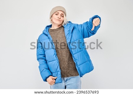 Portrait of blond girl in blue winter clothes showing thumbs down isolated on white studio background. Dislike, wrong choice, negative evaluation concept
