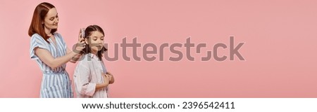 caring mother brushing hair of happy teenage daughter on pink backdrop in studio, horizontal banner