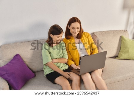 happy woman with teenage daughter watching movie on laptop on couch in living room, quality time