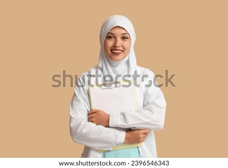 Female Muslim doctor in hijab with clipboard on beige background. World Hijab Day concept