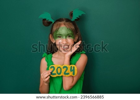 A sweet beautiful girl dressed up as a dragon holds the number 2024 in her hands. The child is inspired by the expectation of the new year