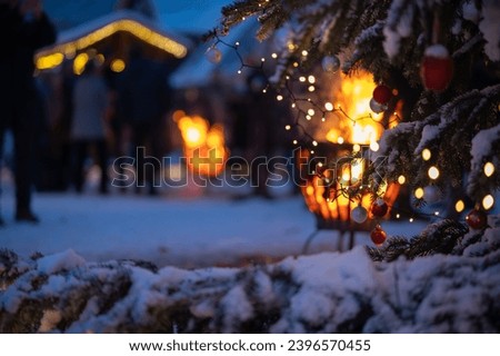 Fir tree branches covered with snow, adorned with sparkling Christmas balls,set against warm glow of a fire pit with delightful bokeh. Scene from Christmas market with snow, Christmas tree, fire bowl.