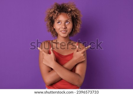 Young happy funny African American woman teen points fingers two hands in different directions paying attention to place for advertising and looking up posing on isolated purple background. Copy space