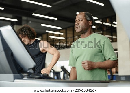 Elderly black man standing on treadmill at gym with caucasian gym goer working out on background