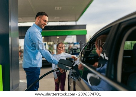 The father pours fuel into the car while his daughter watches him and keeps him company, younger daughter is sitting in the car Royalty-Free Stock Photo #2396594701