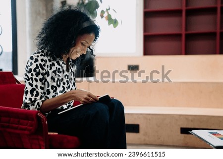 Joyful Black businesswoman in a leopard print blouse using a tablet in a relaxed modern office setting with stylish red furniture Royalty-Free Stock Photo #2396611515
