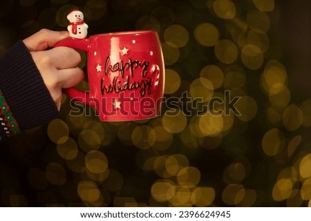 Delicious fresh festive morning coffee in a red cup on the red surface with Christmas tree lights and spruce branches in the background. 