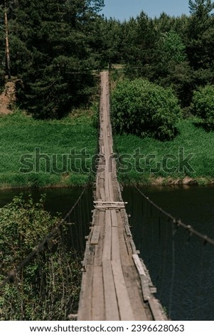 old rusty small wooden plank bridge over river in countryside, sunny summer day, green trees in background