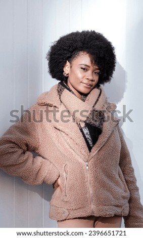 Stylish model posing in front of white wall outdoors. She is smiling and looking at camera wearing a winter coat and scarf. She has a curly black hair and the sun is hitting her face.