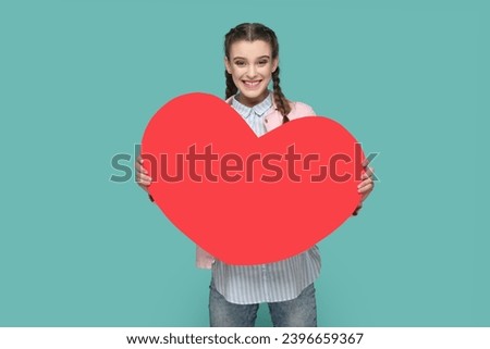 Portrait of happy joyful cheerful teenager girl with braids wearing pink jacket holding big red heart, looking at camera. Indoor studio shot isolated on green background.