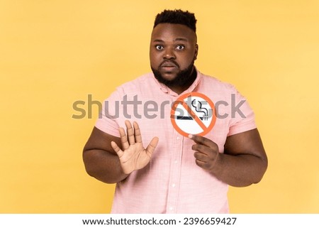 Portrait of young adult bearded man wearing pink shirt holding no smoking sign, showing stop gesture with palm, looking at camera. Indoor studio shot isolated on yellow background.