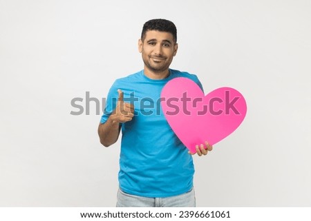 Portrait of joyful romantic unshaven man wearing blue T- shirt standing holding big pink heart and showing thumb up, like gesture. Indoor studio shot isolated on gray background.