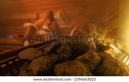Steam rising from sauna stones with blurred people in the background, warm glowing light. relaxing in finnish sauna spa hotel concept image Royalty-Free Stock Photo #2396663023