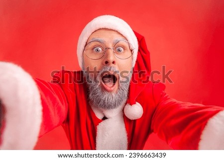 Portrait of elderly man with gray beard in santa claus costume making selfie POV or broadcasting live stream from Christmas party, excited expression. Indoor studio shot isolated on red background.