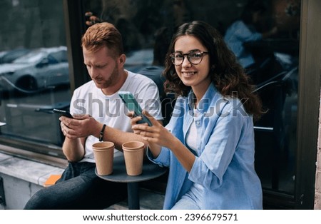 Portrait of happy Caucasian female blogger in optical spectacles for vision correction smiling at camera while millennial boyfriend browsing website on modern smartphone device, technology users Royalty-Free Stock Photo #2396679571