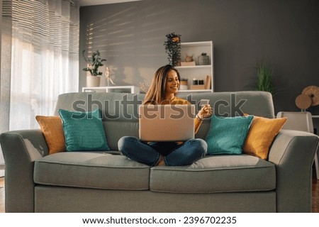 A cheerful brunette is sitting at her comfortable apartment with a laptop in her lap and credit card in the hand and purchasing online on the internet. A woman is using laptop for ordering online. Royalty-Free Stock Photo #2396702235