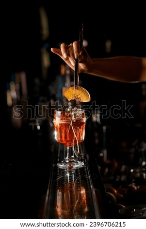 Bartender woman adding slice of peach with tongs to cocktail glass with alcoholic drink at bar counter. Bartender creates an alcoholic drink for women. Royalty-Free Stock Photo #2396706215