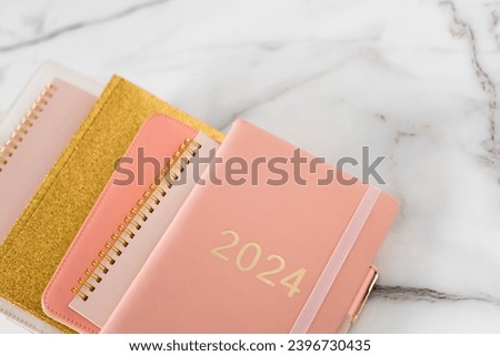 Pink coral colored diary for the year 2024 and many other diaries, pen, marble background. New year planning concept. Minimalistic workstation. Copy space.