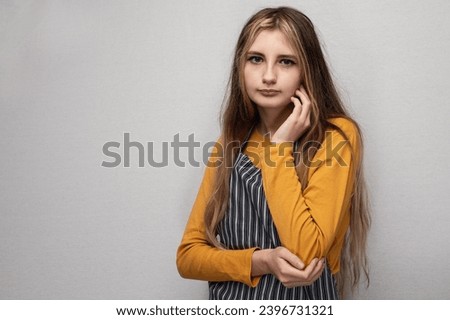 Portrait of young teenage girl in yellow shirt and classic black and white stripe apron on light grey background. Calm face expression, long hair. Home kitchen or art work out wear